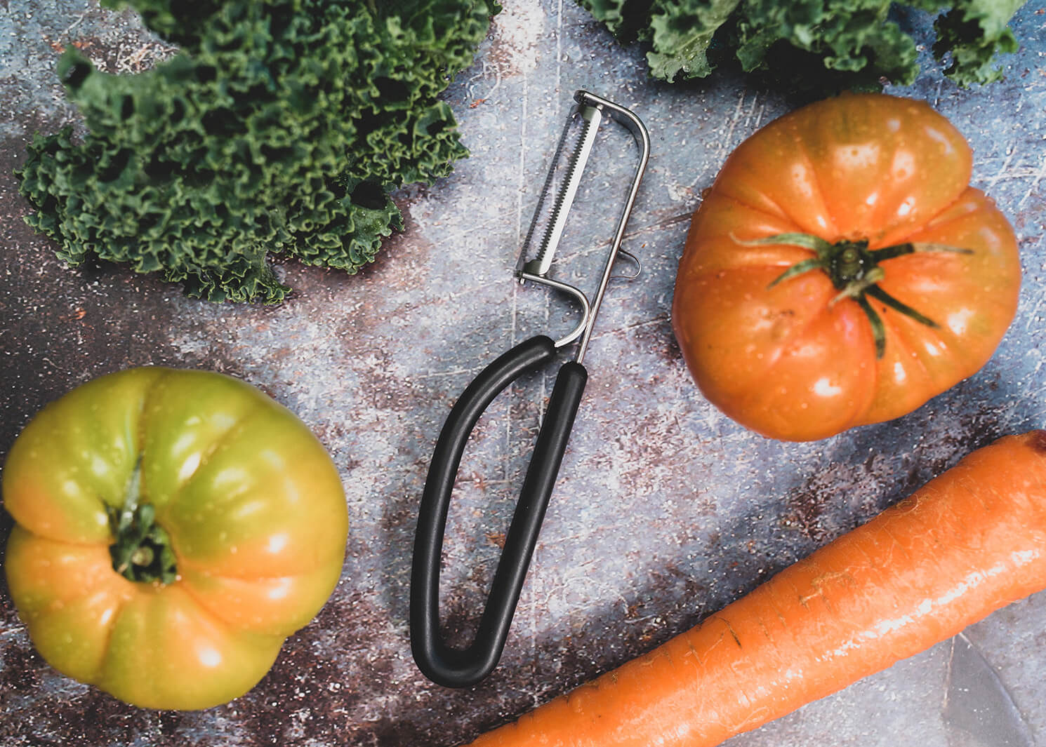 Spartan peeler with Japanese-steel blade and black ergonomic handle lays on cutting board with field tomatoes and a large carrot